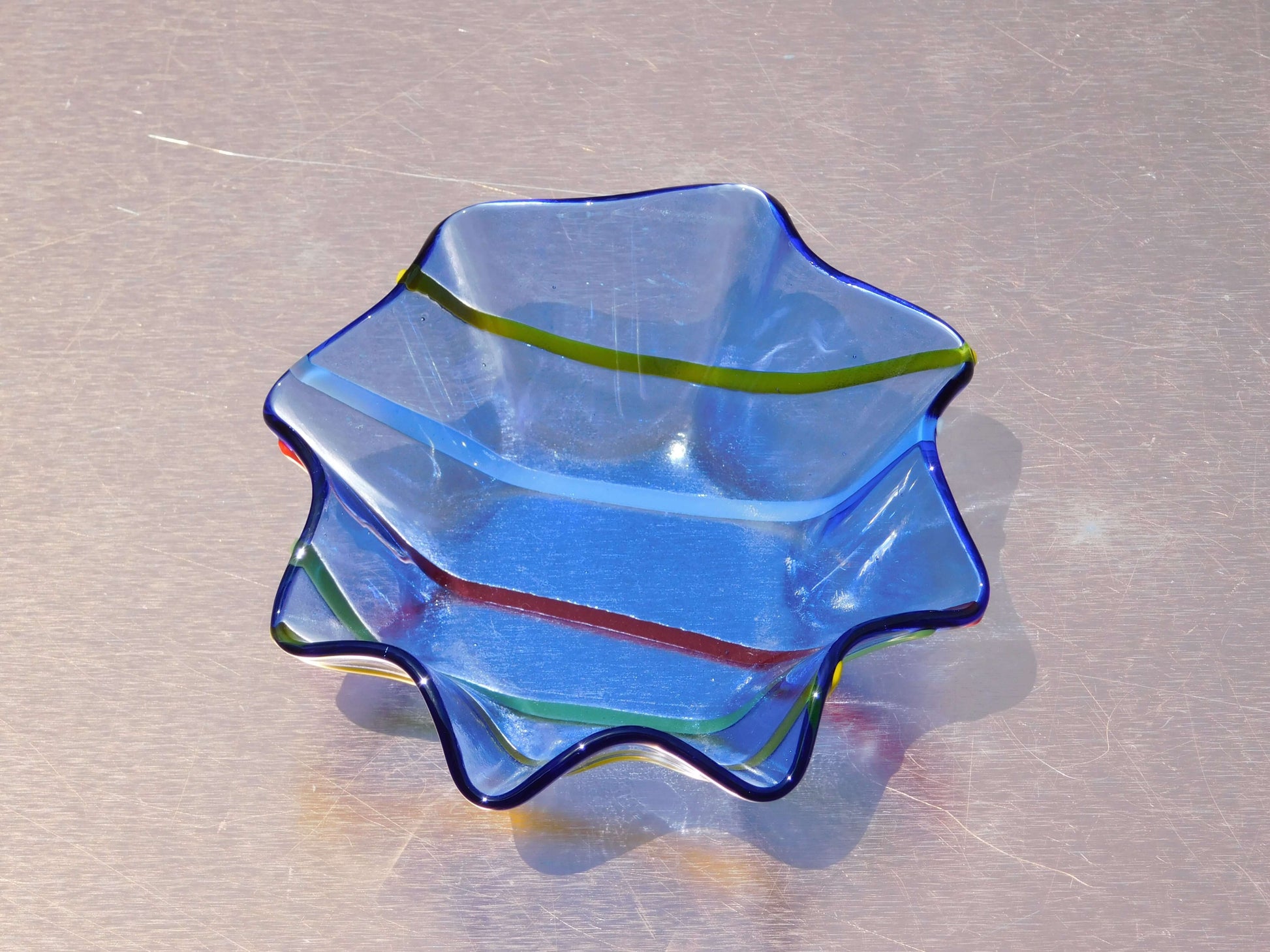 A blue glass decorative trinket bowl with colorful stripes and ruffled edges, displayed against a reflective surface.